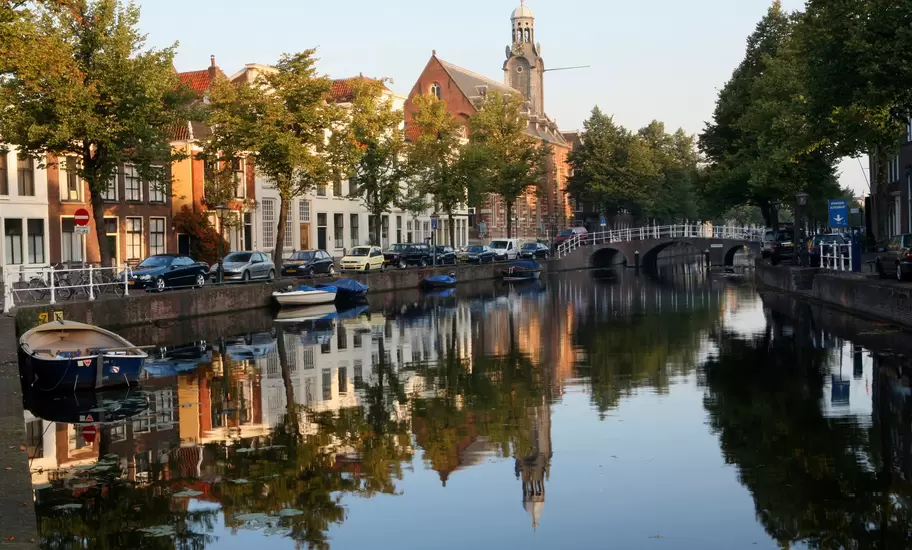 Leiden canal lined with trees, gabled houses and parked cars, a brick building with cupola tower reflected in the water