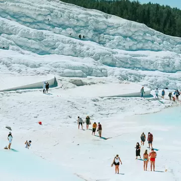 Visitors walk barefoot across white travertine terraces and pale blue thermal pools below a pine-covered hill at Pamukkale
