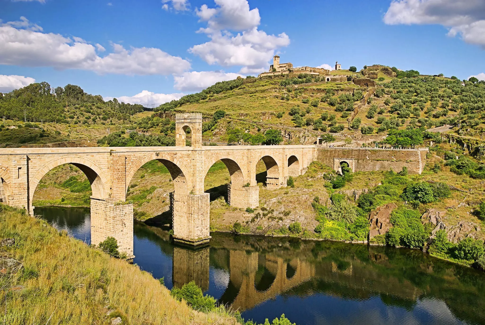 A stone bridge with an arch in the middle of it spans over a body of water between two pieces of lush, green land