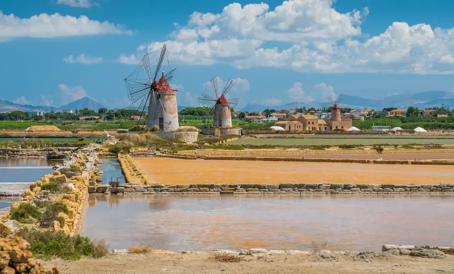 Red-capped windmills behind stone-edged salt pans in pink and ochre at the Stagnone lagoon, low town and mountains beyond