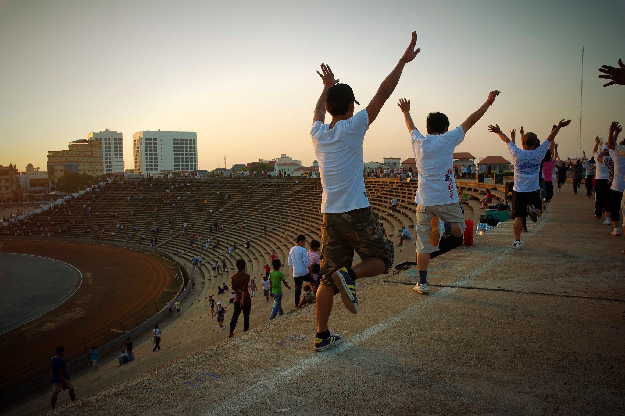 People exercising and standing on the stairs and walkways of an open-air stadium in Cambodia in warm light.