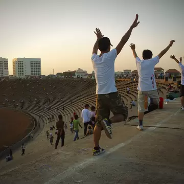 People exercising and standing on the stairs and walkways of an open-air stadium in Cambodia in warm light.