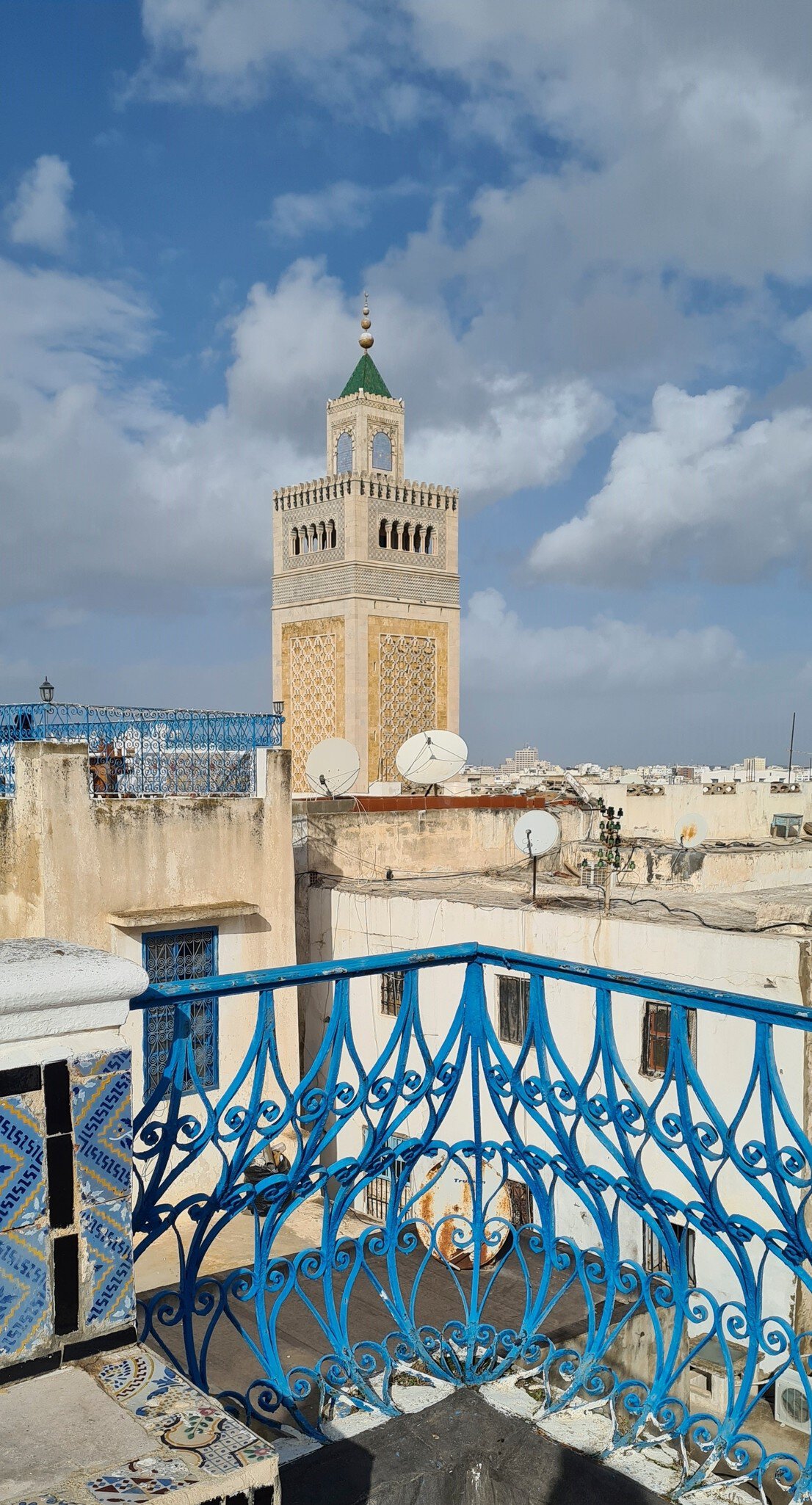 Tall carved stone tower with a green pyramid top seen over flat roofs and a blue wrought-iron balcony rail.