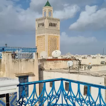 Tall carved stone tower with a green pyramid top seen over flat roofs and a blue wrought-iron balcony rail.
