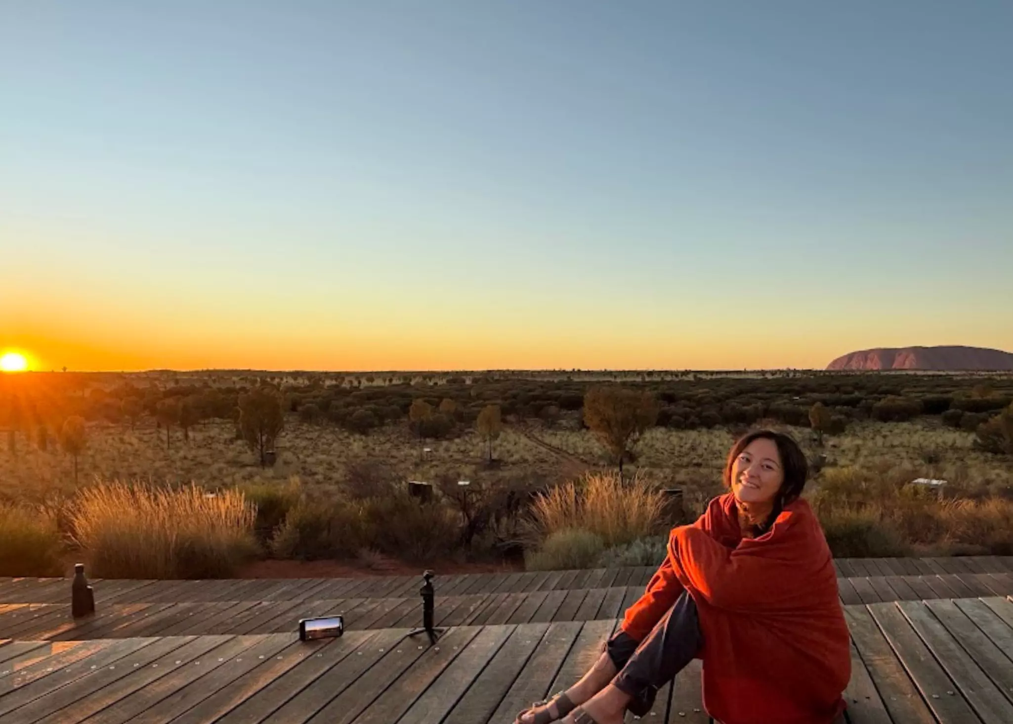 A woman sits on some decking near a vast desert area as the sun comes up over the horizon.