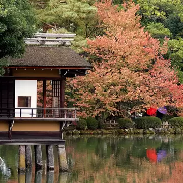 Wooden teahouse on stilts over a pond in Kenroku-en Garden, Kanazawa, with red autumn foliage beyond