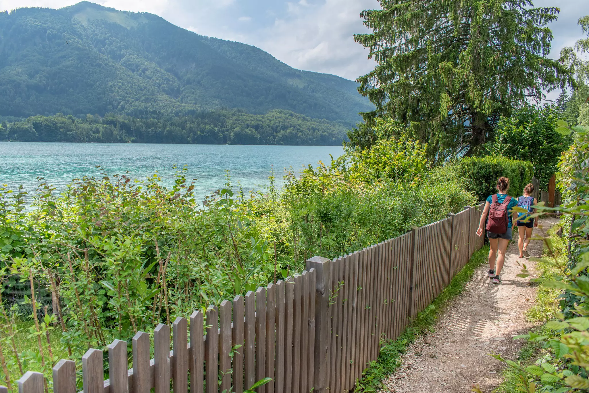 Two hikers on a dirt path alongside a blue lake.