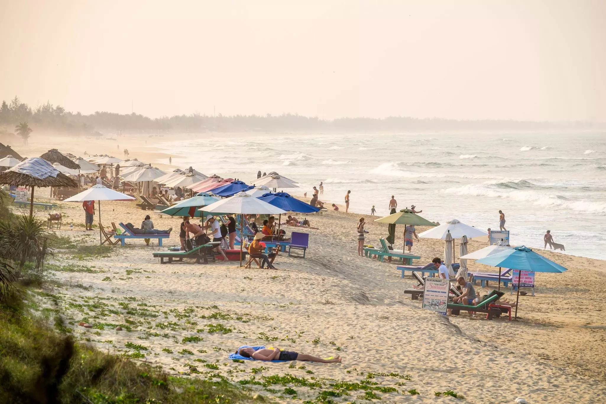 People under sunshades on a busy beach with waves crashing on the shore.