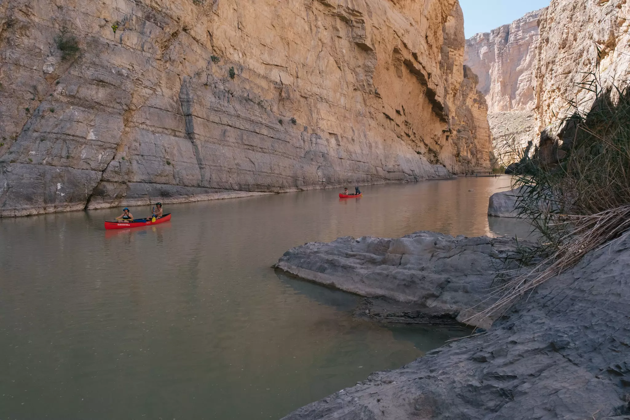 Two red canoes paddle on a narrow river running through a deep, shadowy canyon.