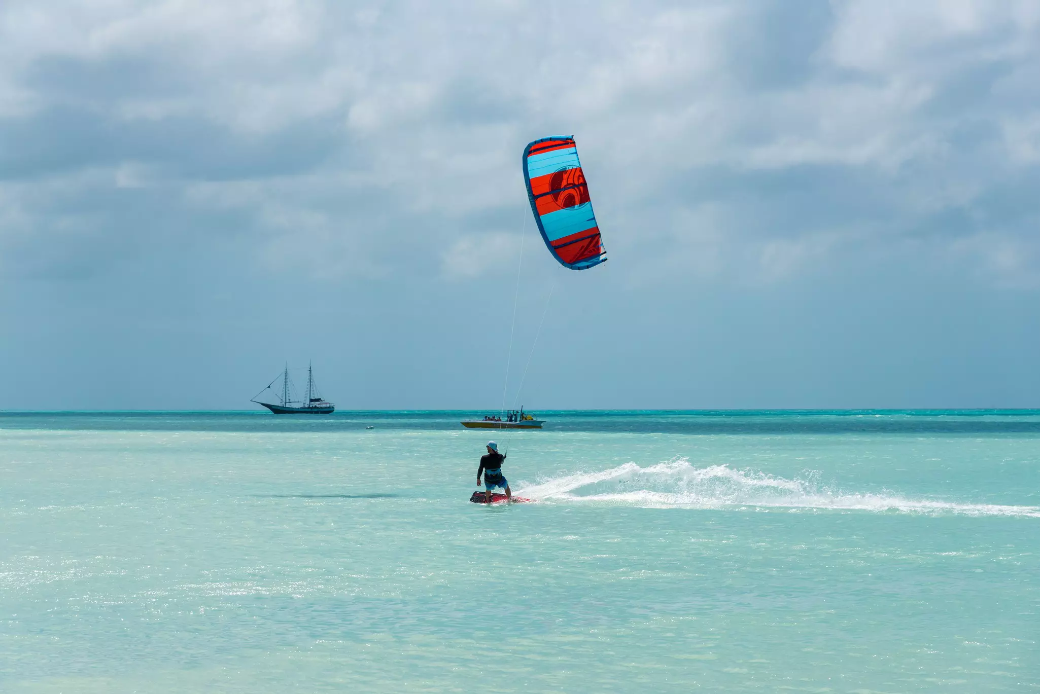 A person on a kiteboard in light blue water with a power boat and sailbot in the distance on a cloudy day.
