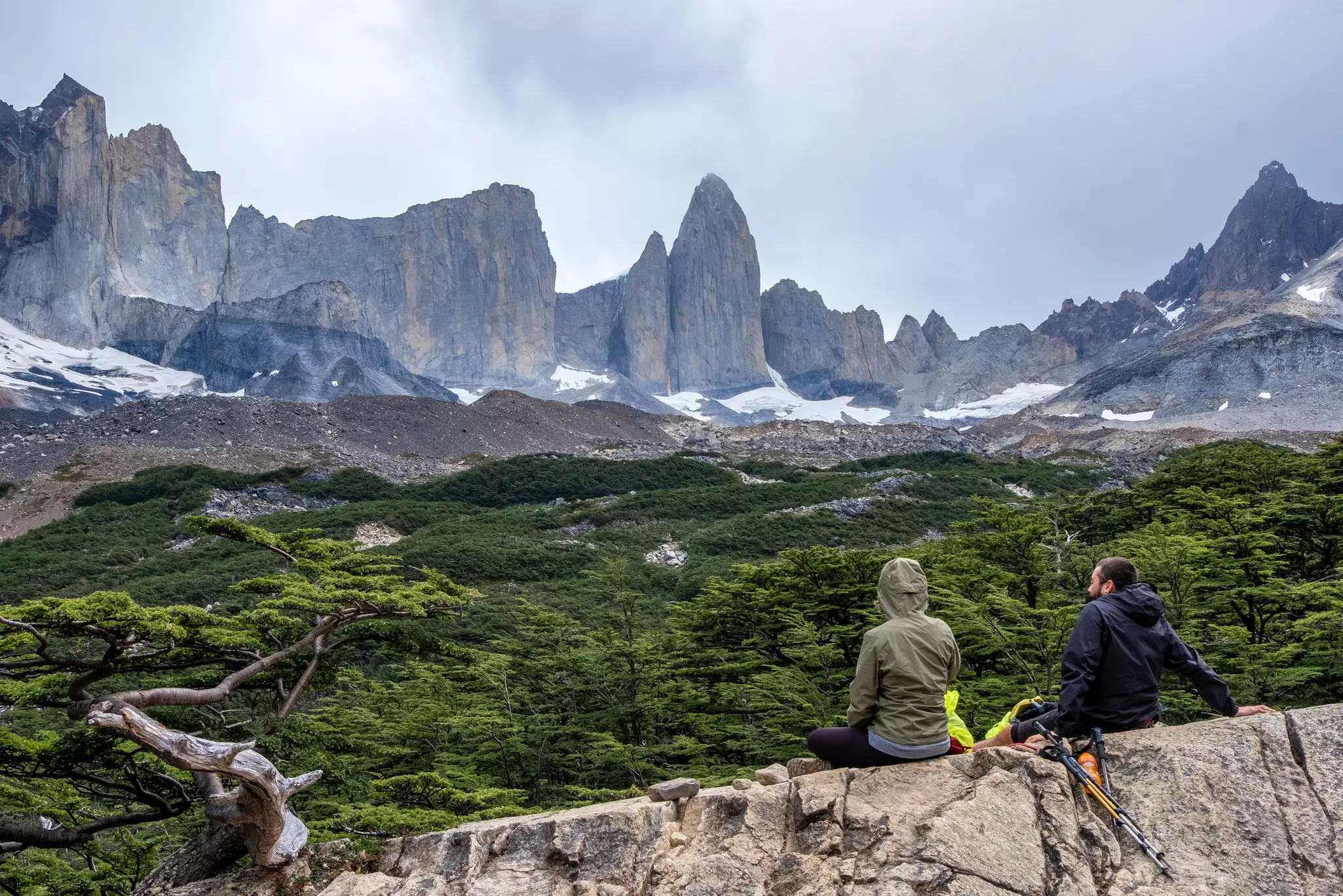 Two hikers sit on a rock looking at craggy mountains across a valley.