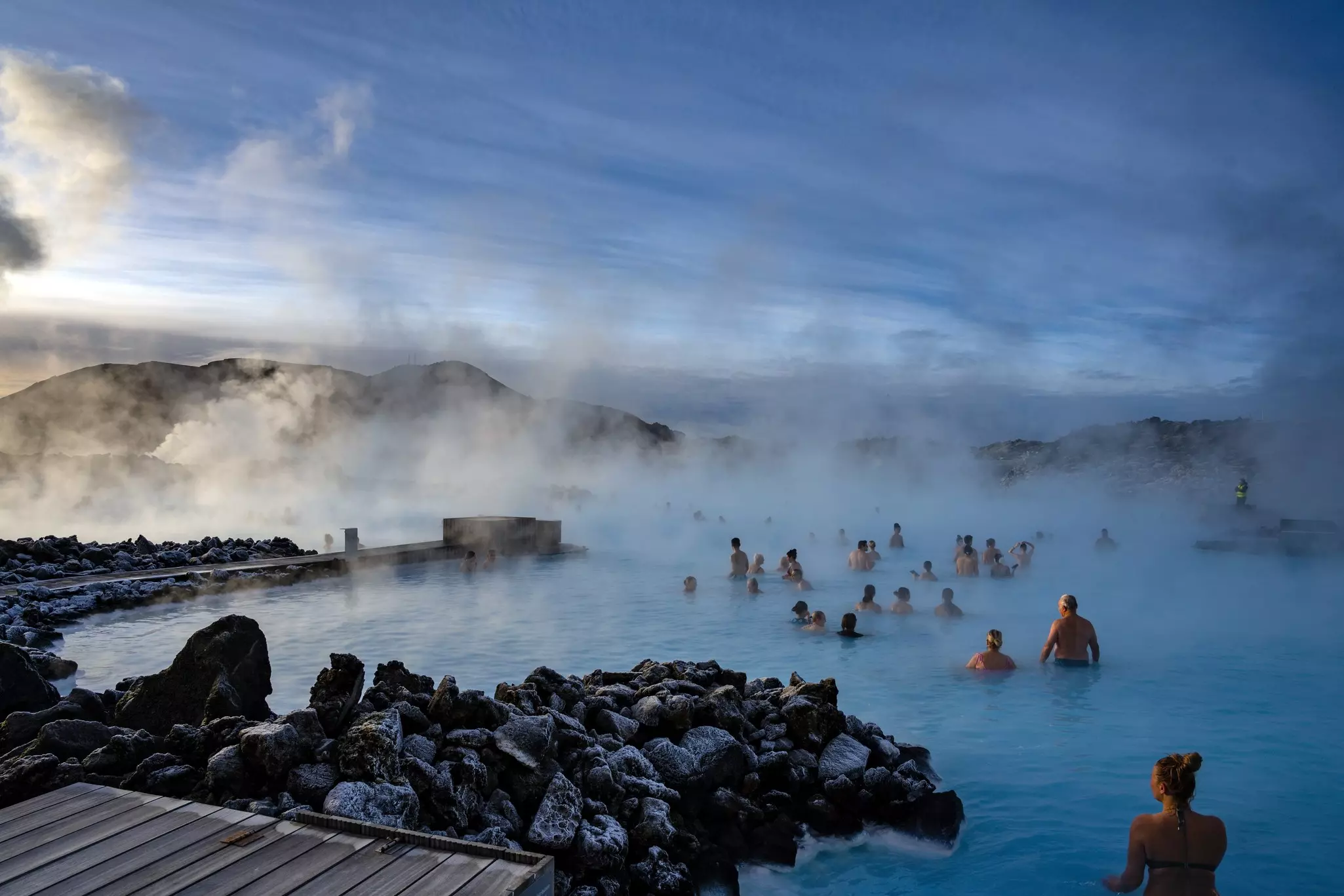 People swimming in a geothermal pool at dusk with steam clouds rising from the deep blue water.