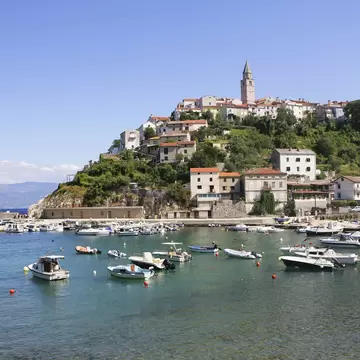 Small motorboats moored in a calm bay below stone houses and a bell tower on a green hillside at Vrbnik.
