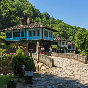 Bright blue timber house with a stone-slab roof at Etâr, visitors crossing a flagstone bridge below forested hills