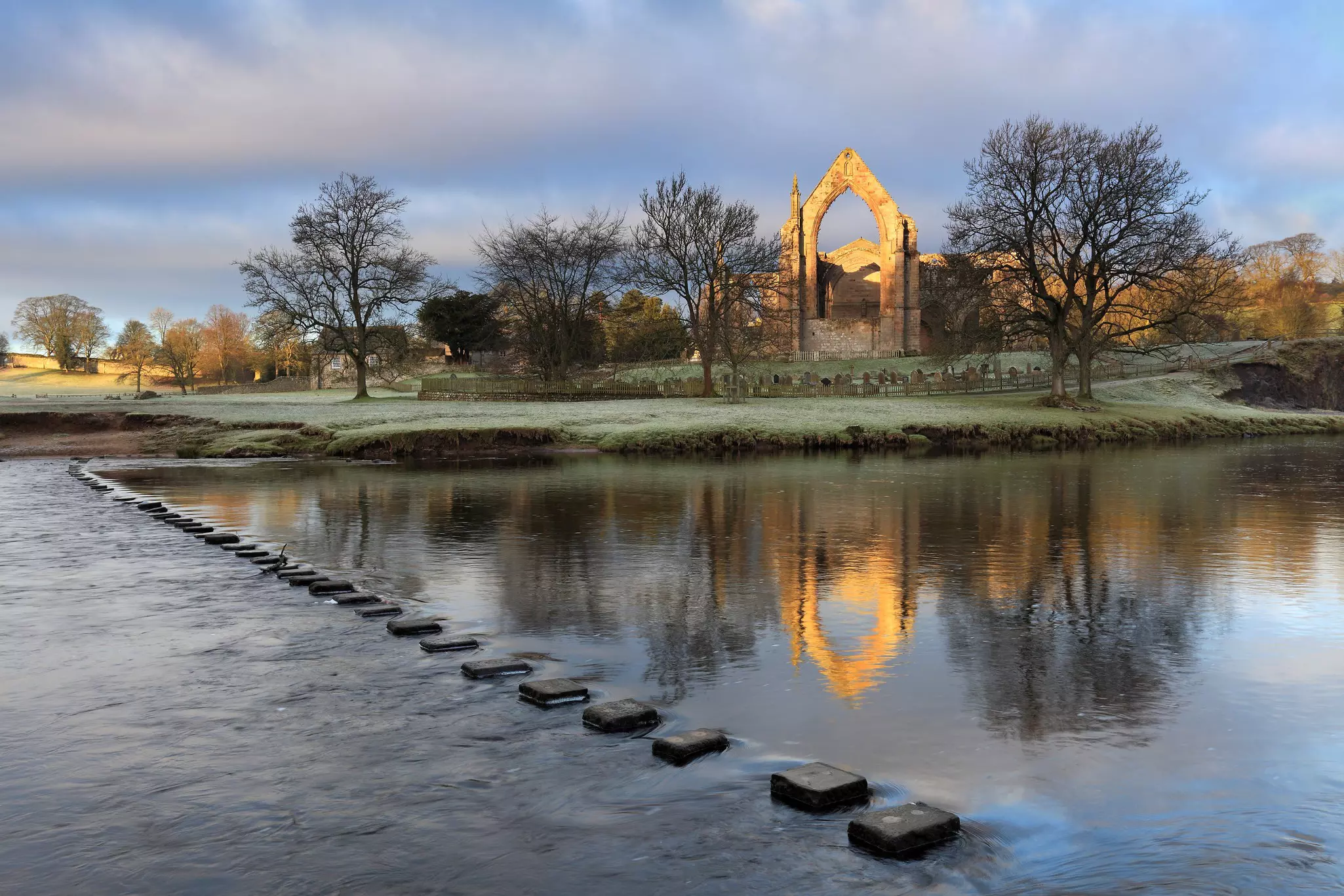 A view of Bolton Abbey, as seen from the opposite bank of the River Wharfe. A series of stepping stones span the river, while the grand abbey is flanked by woodland.