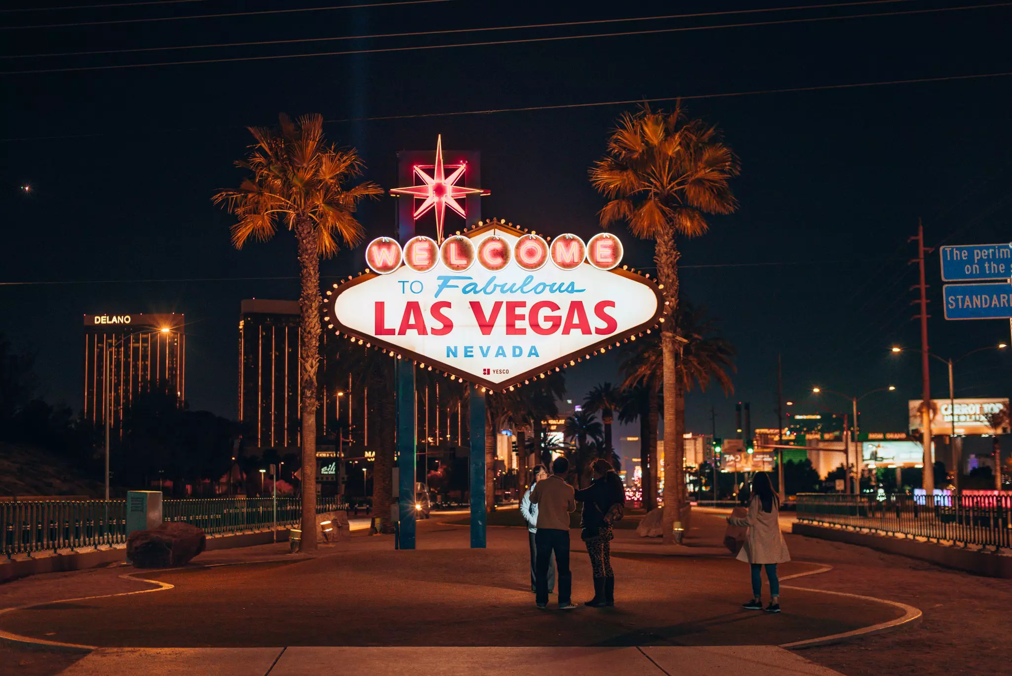 A neon side reading Welcome to Fabulous Las Vegas. It is night time and there are palm trees, buildings and people nearby.