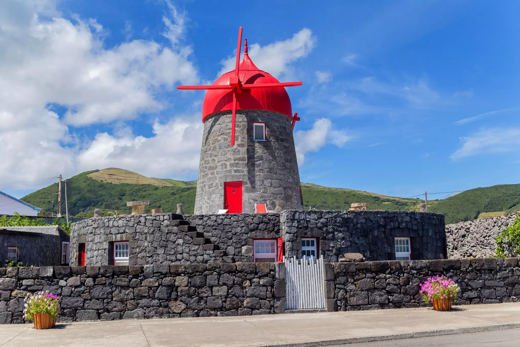 Stone windmill with red roof and red blades surrounded by a low, circular stone building with green hills in the background on a sunny day.