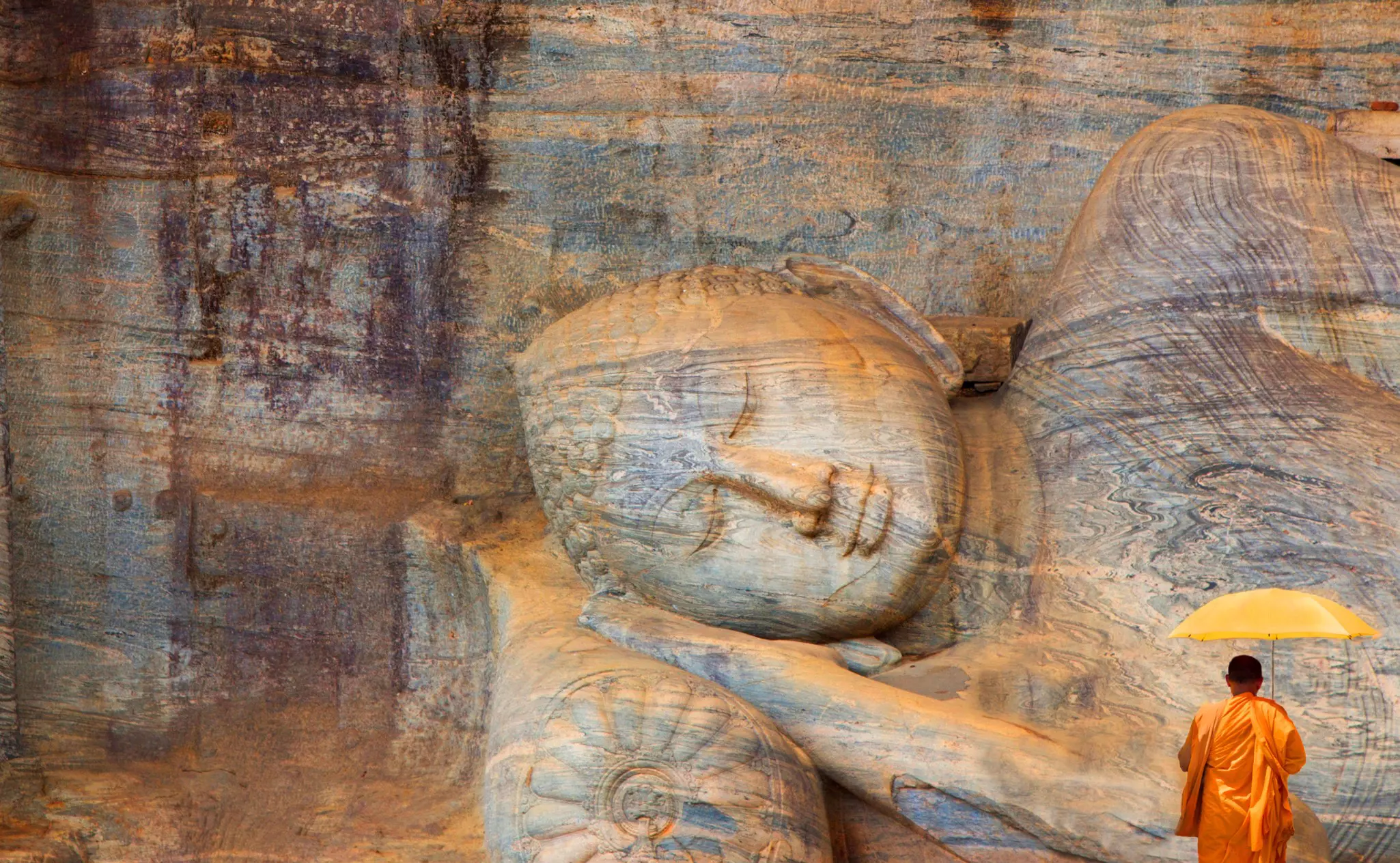 A monk in orange robes with an umbrella pays respect to a statue of the Buddha at Polonnaruwa, Sri Lanka. 