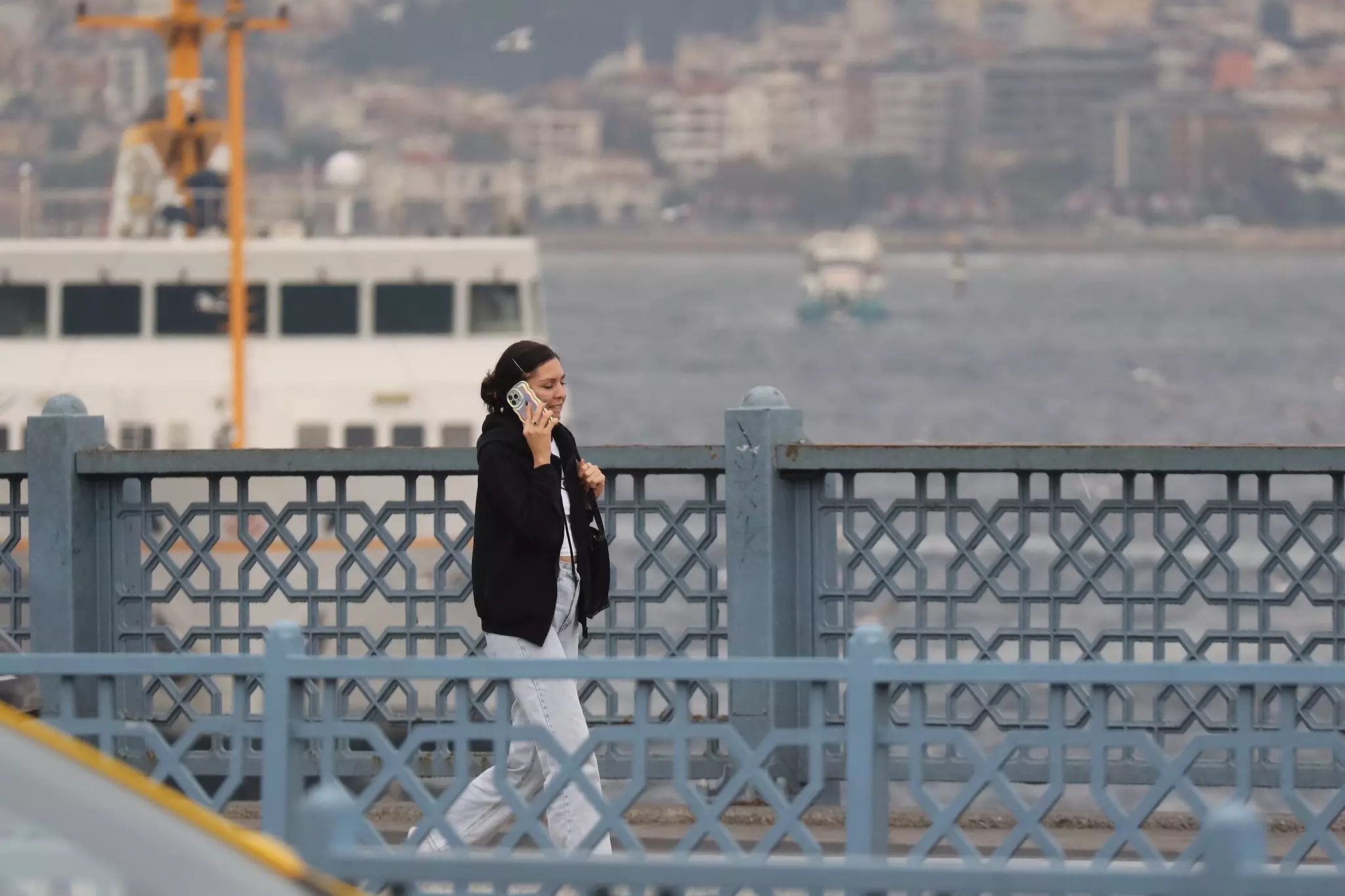 A woman talks on her phone while walking on a bridge with a ferry in the background,
