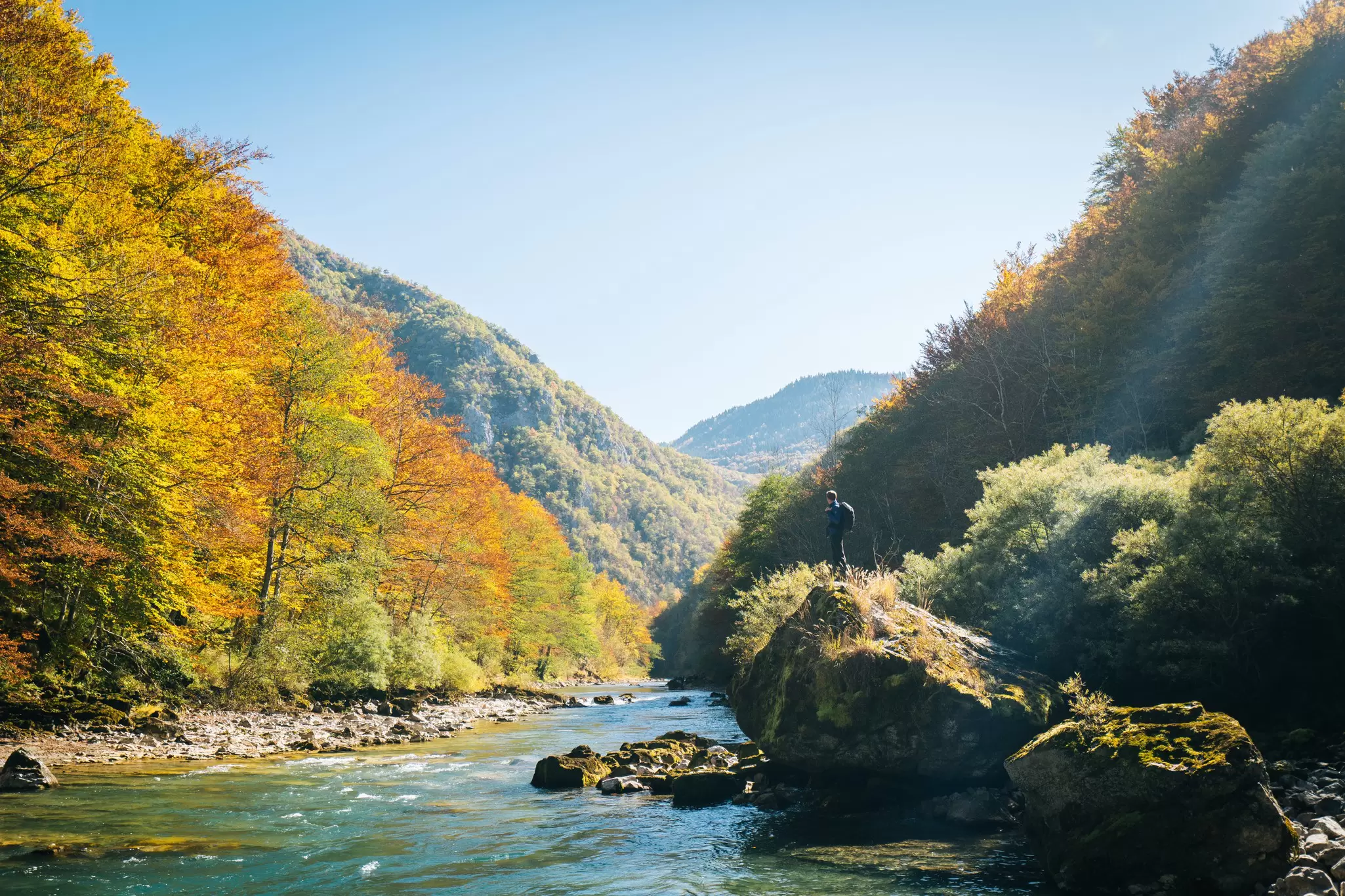 A hiker pauses at a viewpoint to look out across a river towards hillsides covered in golden trees in Montenegro