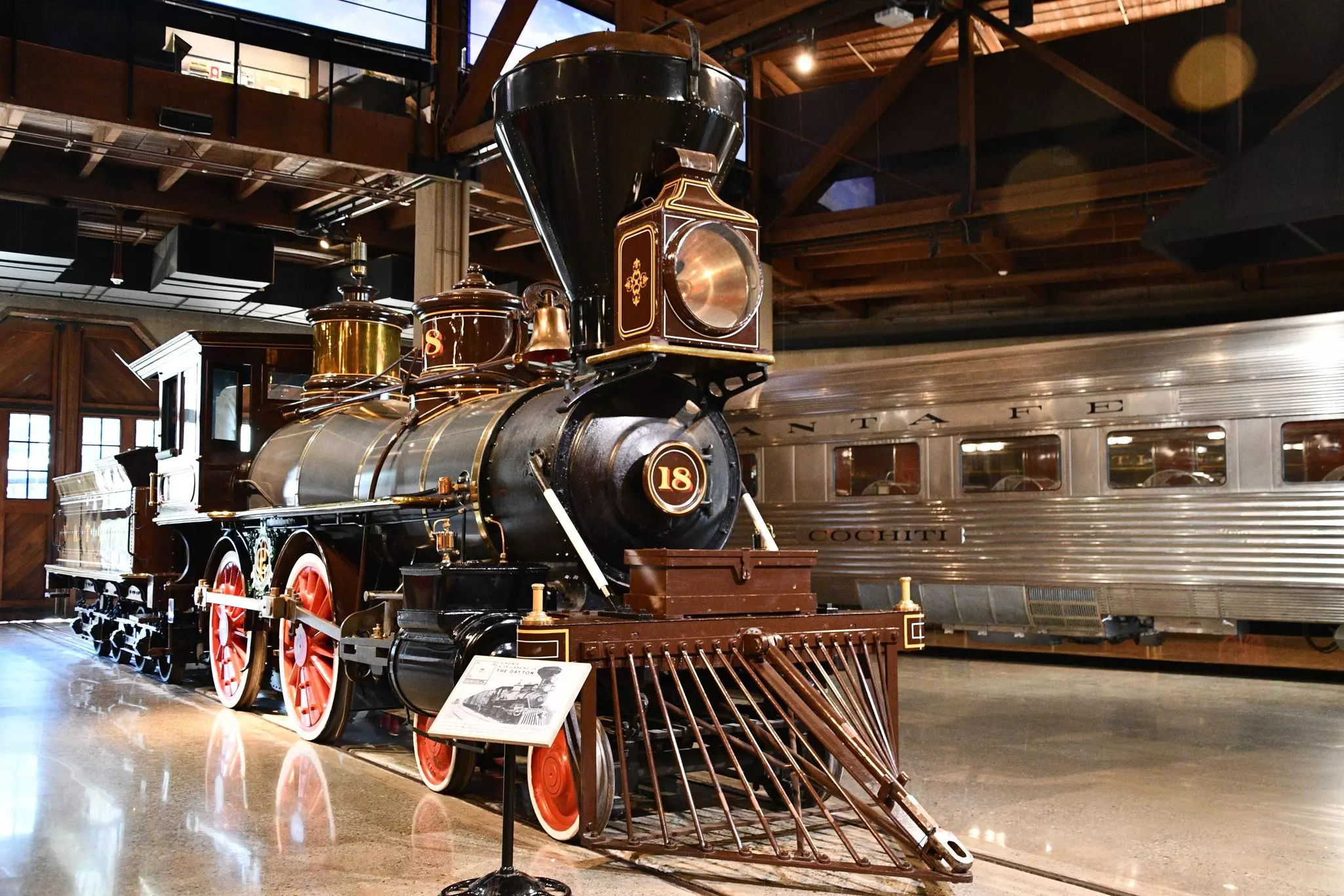 Interior of museum with historic large, black train and silver, more modern train to the right.