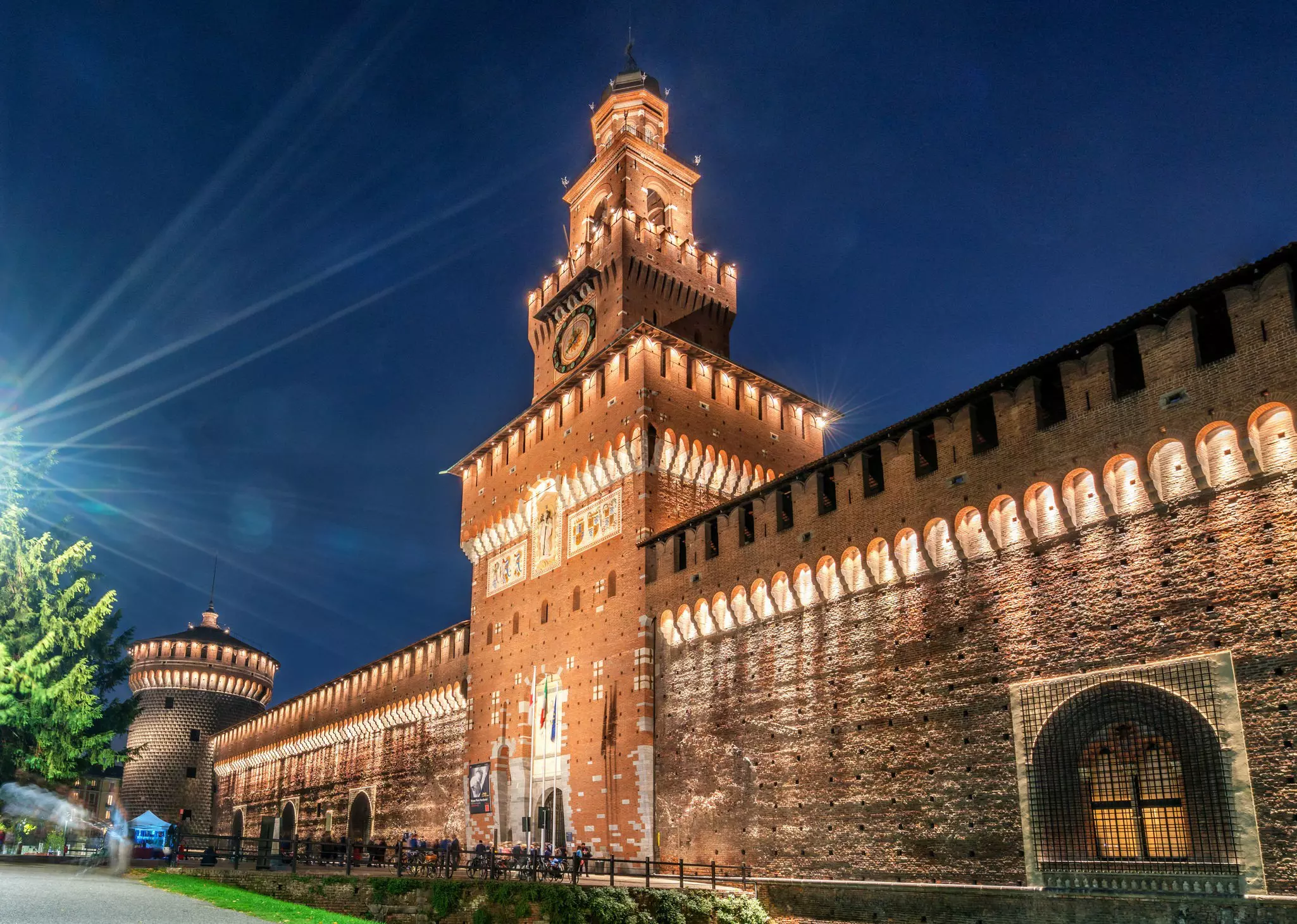 The huge wall of a castle, with a central tower and crenellations, is illuminated at night.