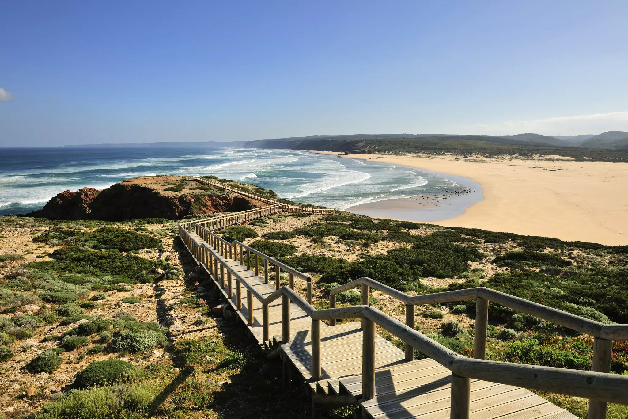 Long wooden walkway leading down to a low cliff overlooking a curved sandy coastline with the ocean beyond on a sunny day.