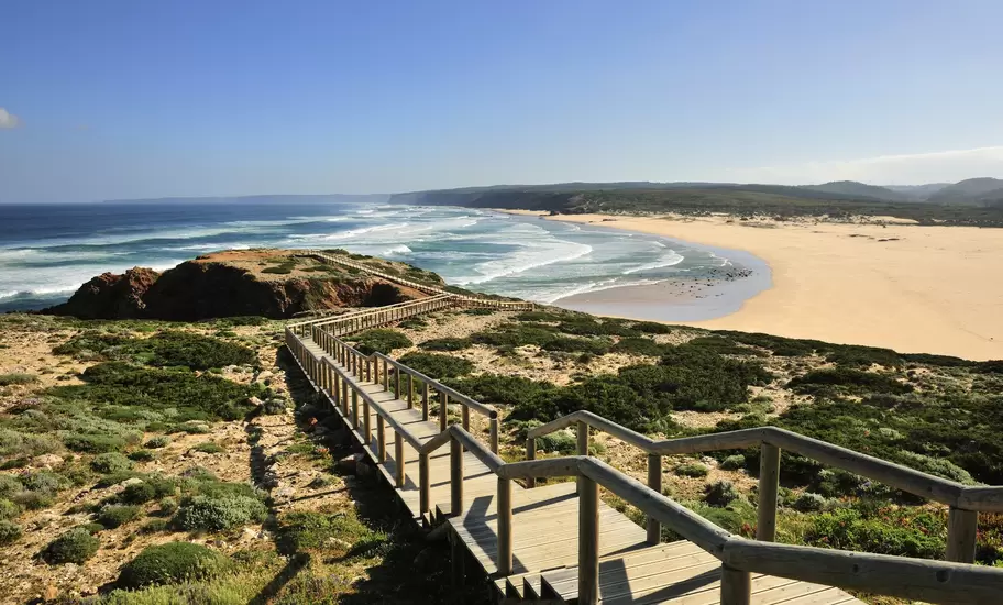 Long wooden walkway leading down to a low cliff overlooking a curved sandy coastline with the ocean beyond on a sunny day.