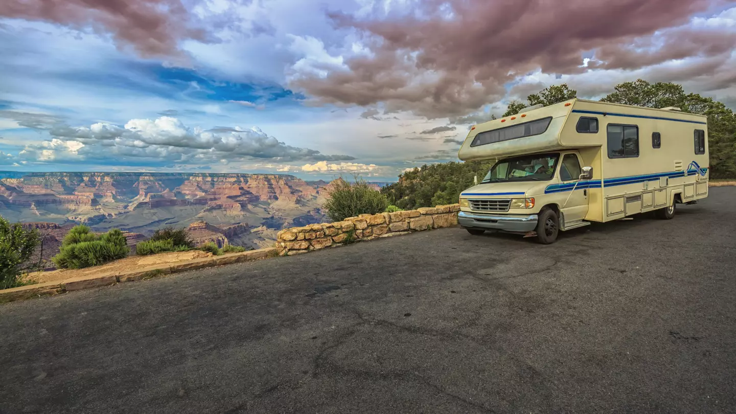 An RV stops on a blacktop road by a low stone wall overlooking the Grand Canyon at sunset.