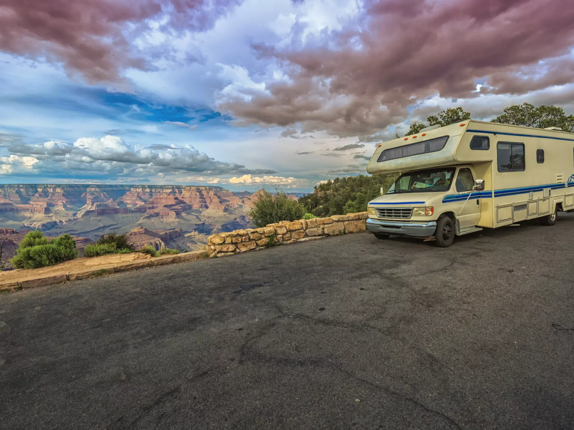 An RV stops on a blacktop road by a low stone wall overlooking the Grand Canyon at sunset.