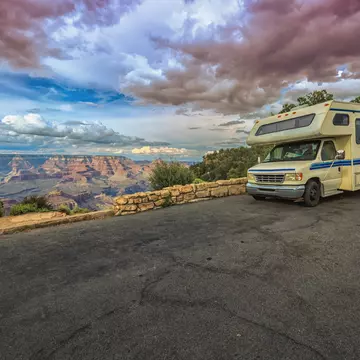 An RV stops on a blacktop road by a low stone wall overlooking the Grand Canyon at sunset.
