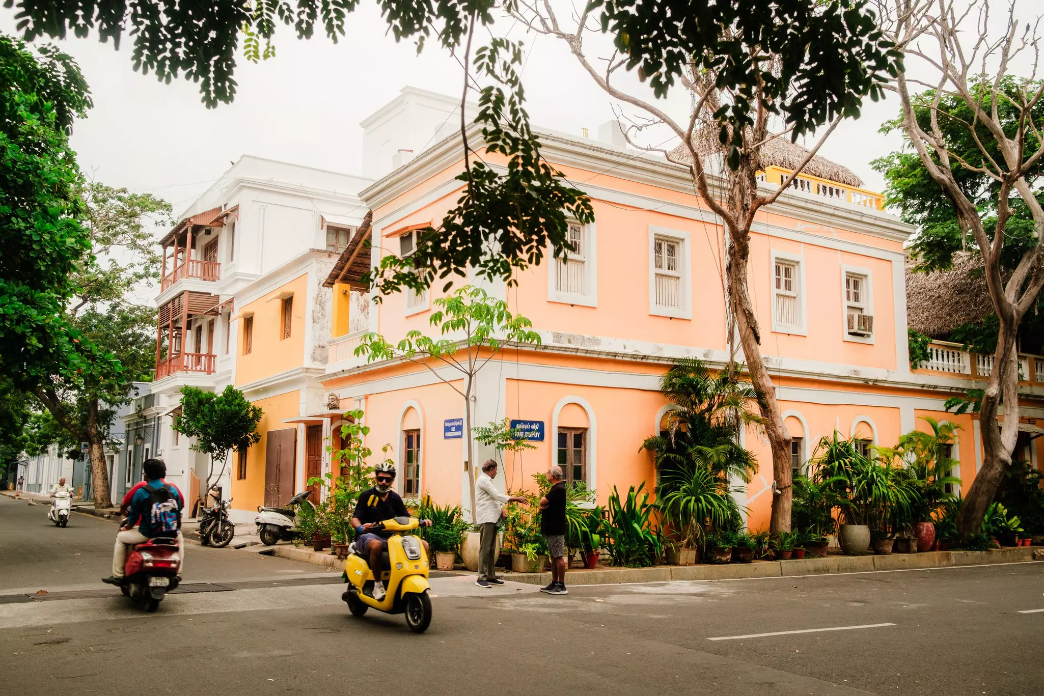 Two men chat on the corner of a street as people pass on motorcycles
