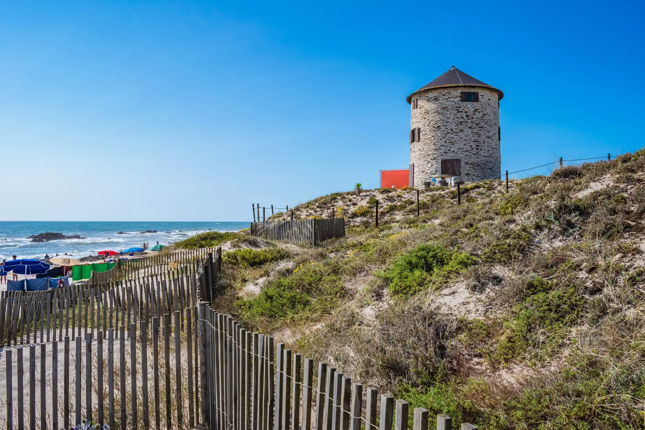 A stone windmill sits at the top of dunes by the ocean in Portugal. A wooden fence is in the foreground, and beach umbrellas are in the distance.