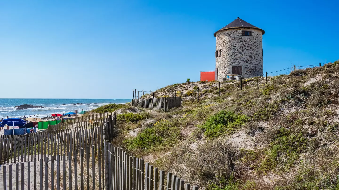 A stone windmill sits at the top of dunes by the ocean in Portugal. A wooden fence is in the foreground, and beach umbrellas are in the distance.