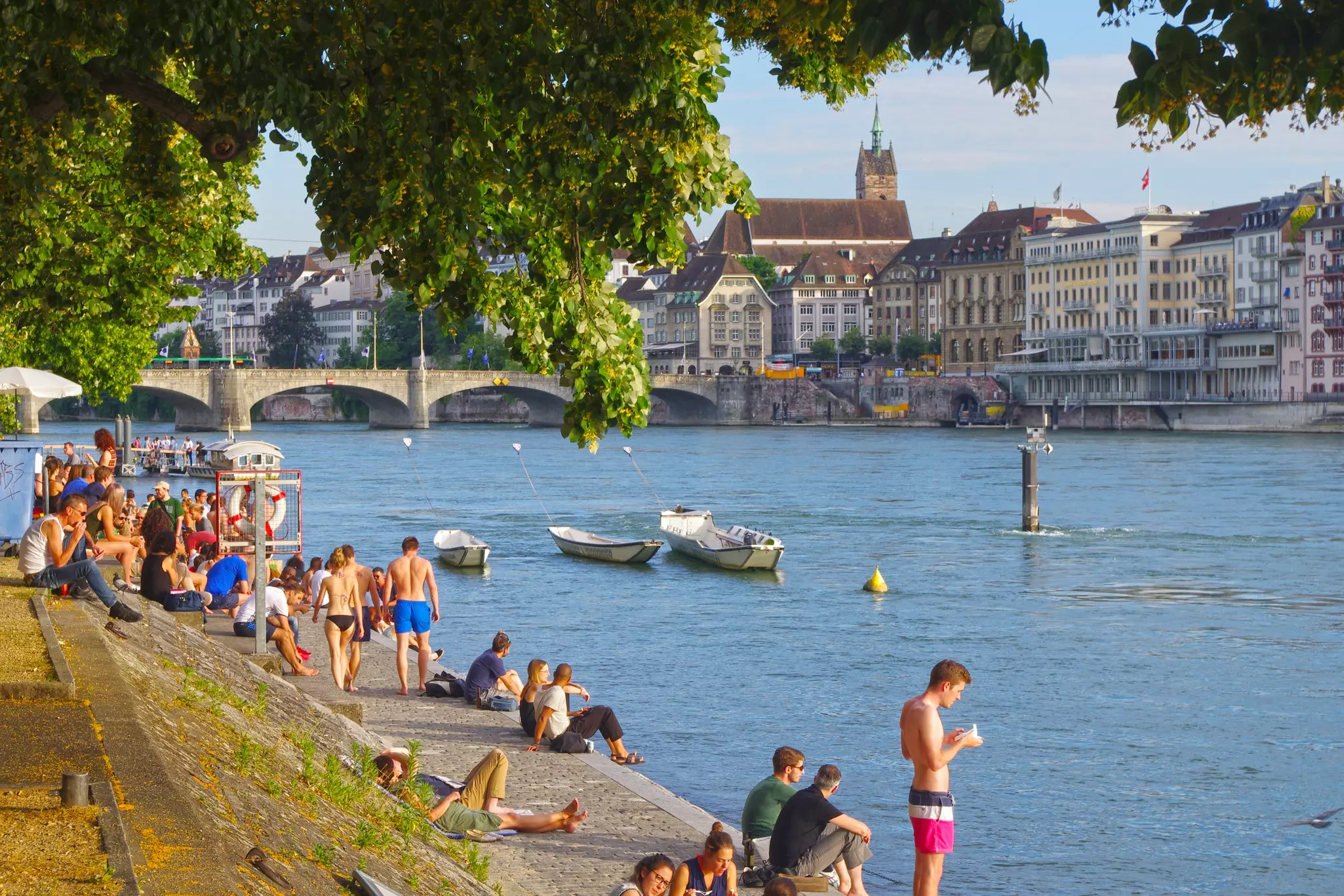 Swimmers walk along the riverbank of a historic city on a summer day.