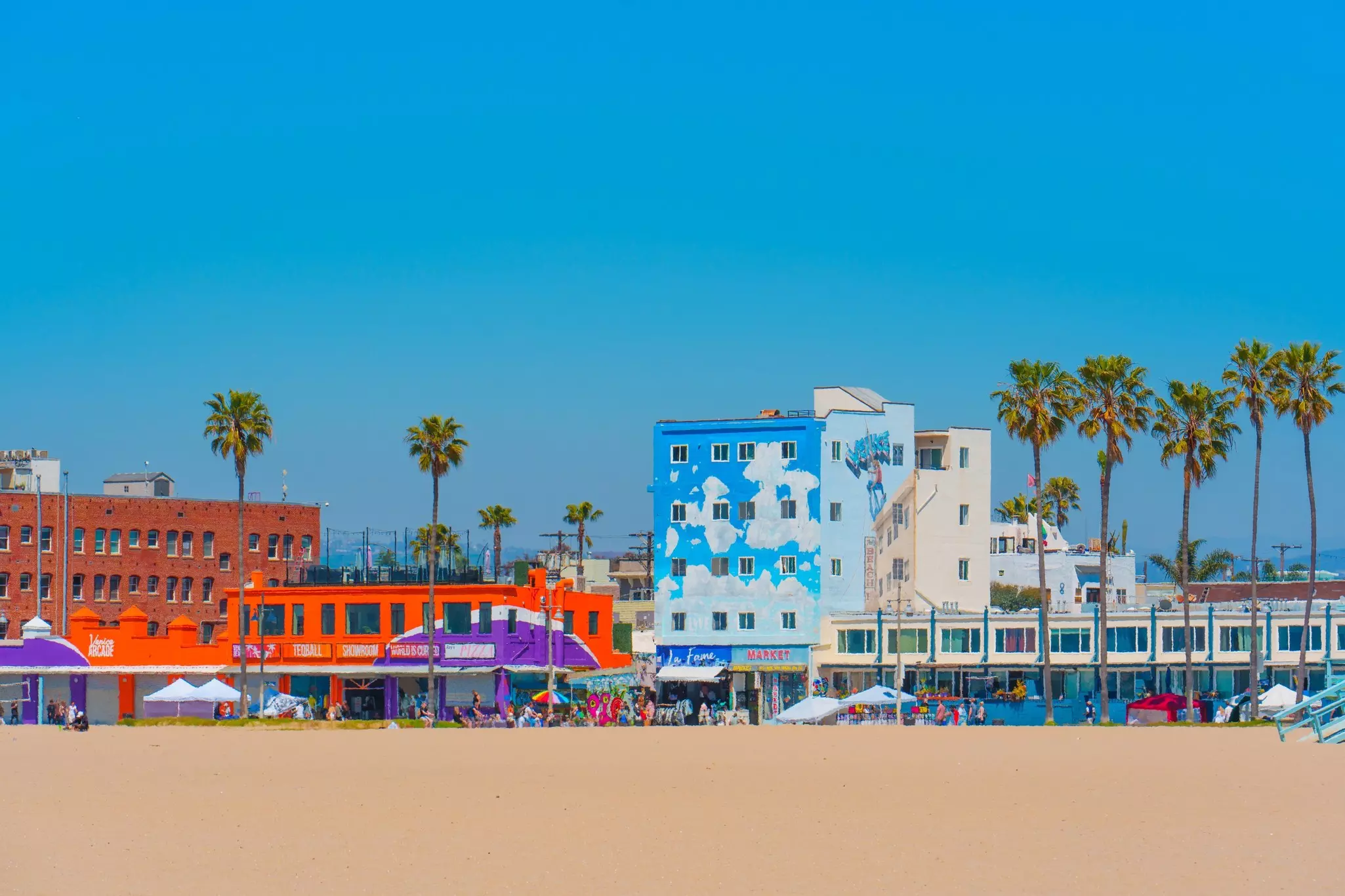 View from Venice Beach with sand in the foreground and colorful buildings and market stalls along the beach line against a clear blue sky.