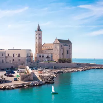 Pale stone cathedral with a tall bell tower on a seafront, turquoise water in front and a small white sailboat.