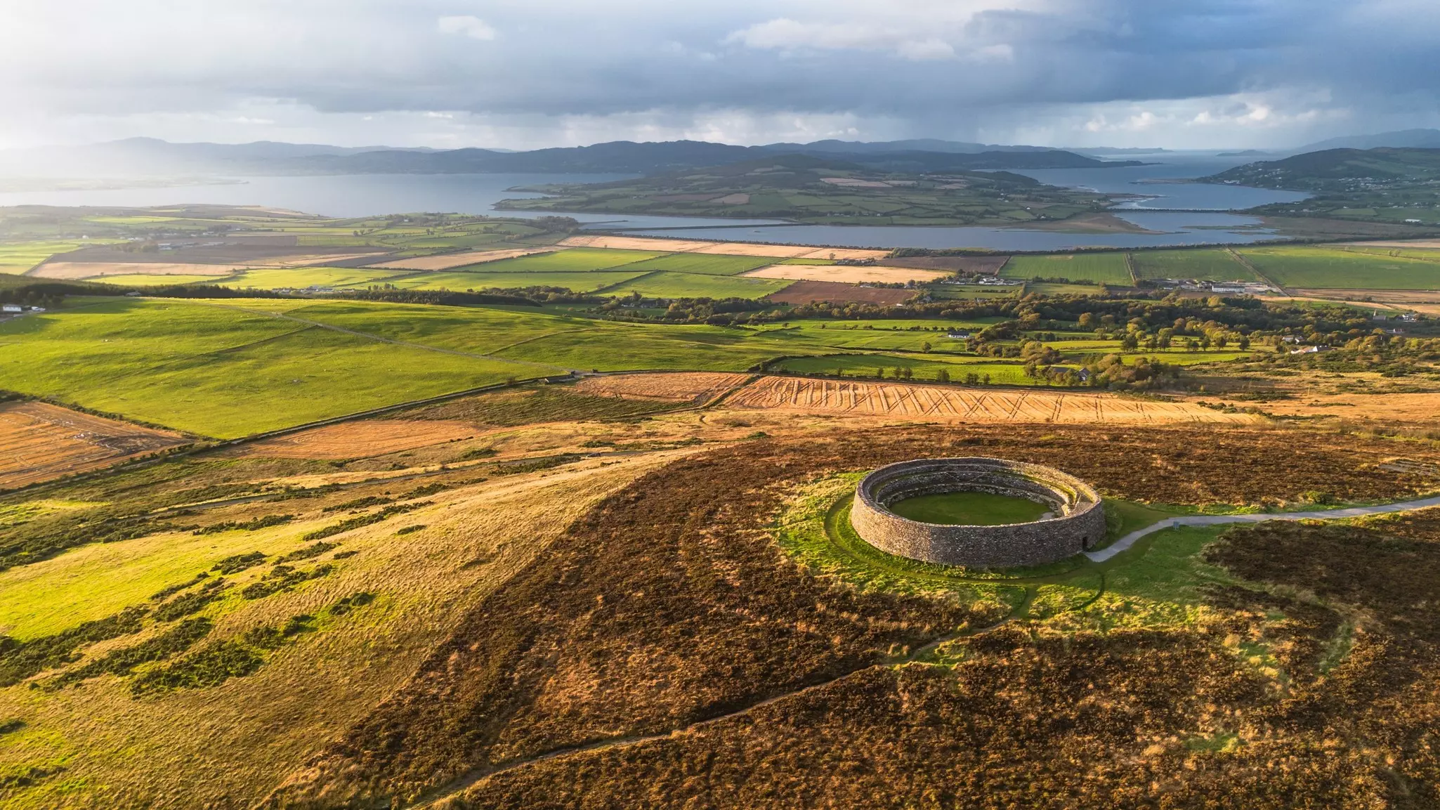 Captivating aerial landscape featuring the historic Grianán of Aileach, a unique circular structure surrounded by vibrant green and brown fields under the golden light of sunset
