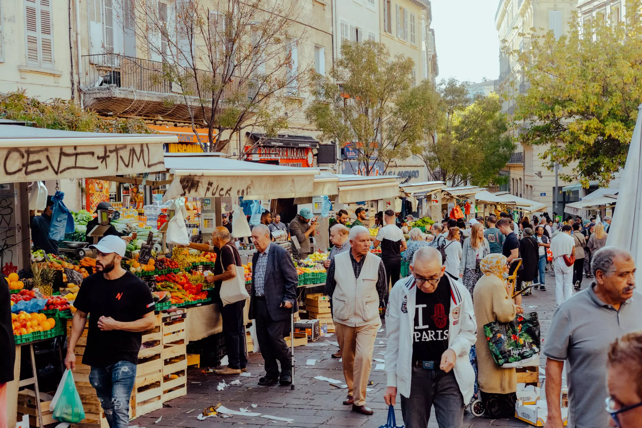 People walk through an outdoor market; stands under stands have piles of brightly colored produce.