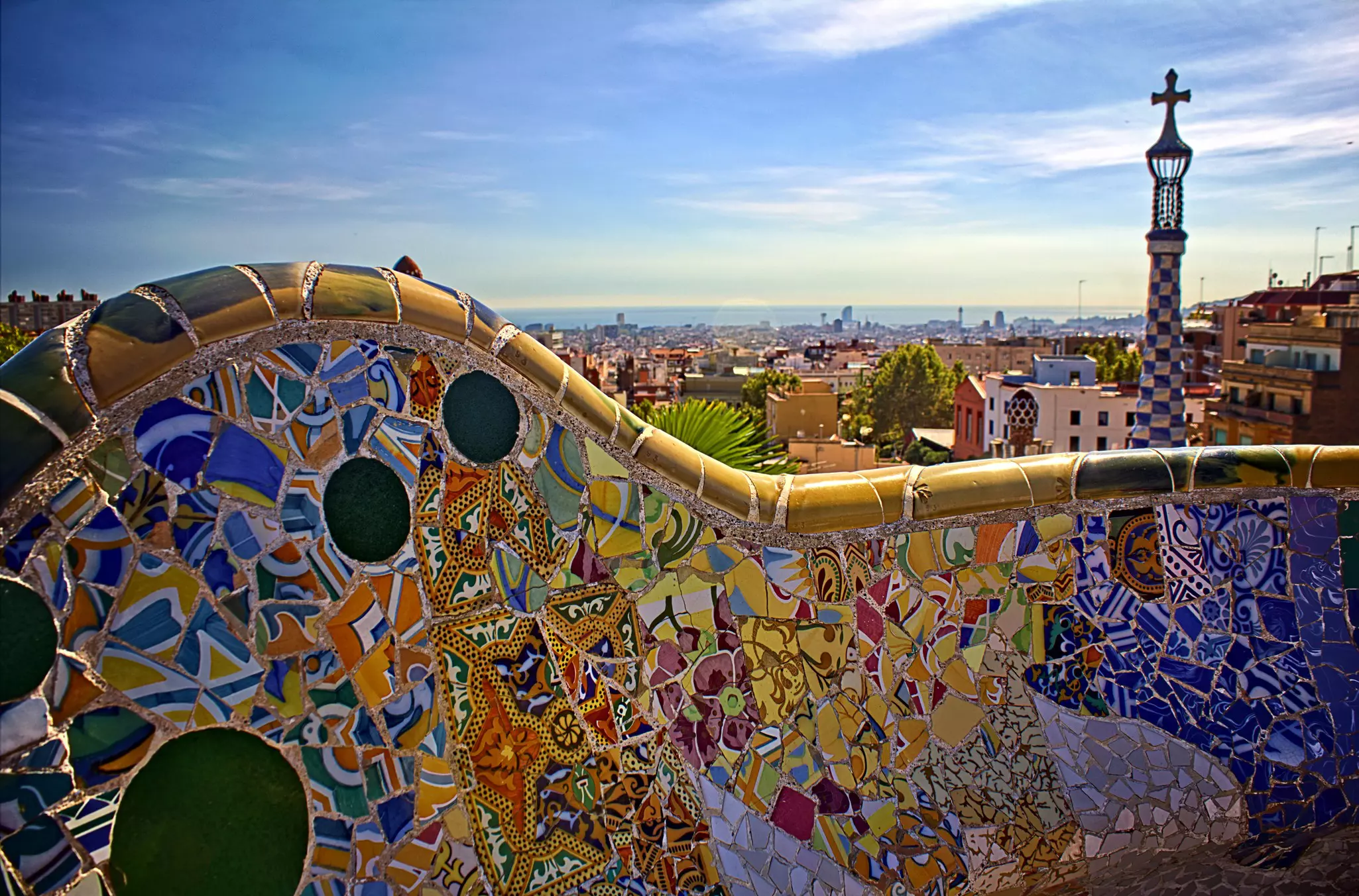 Multicolored mosaic designs on a bench at a park in Barcelona, Spain; a checkerboard tower with a cross is visible in the background.