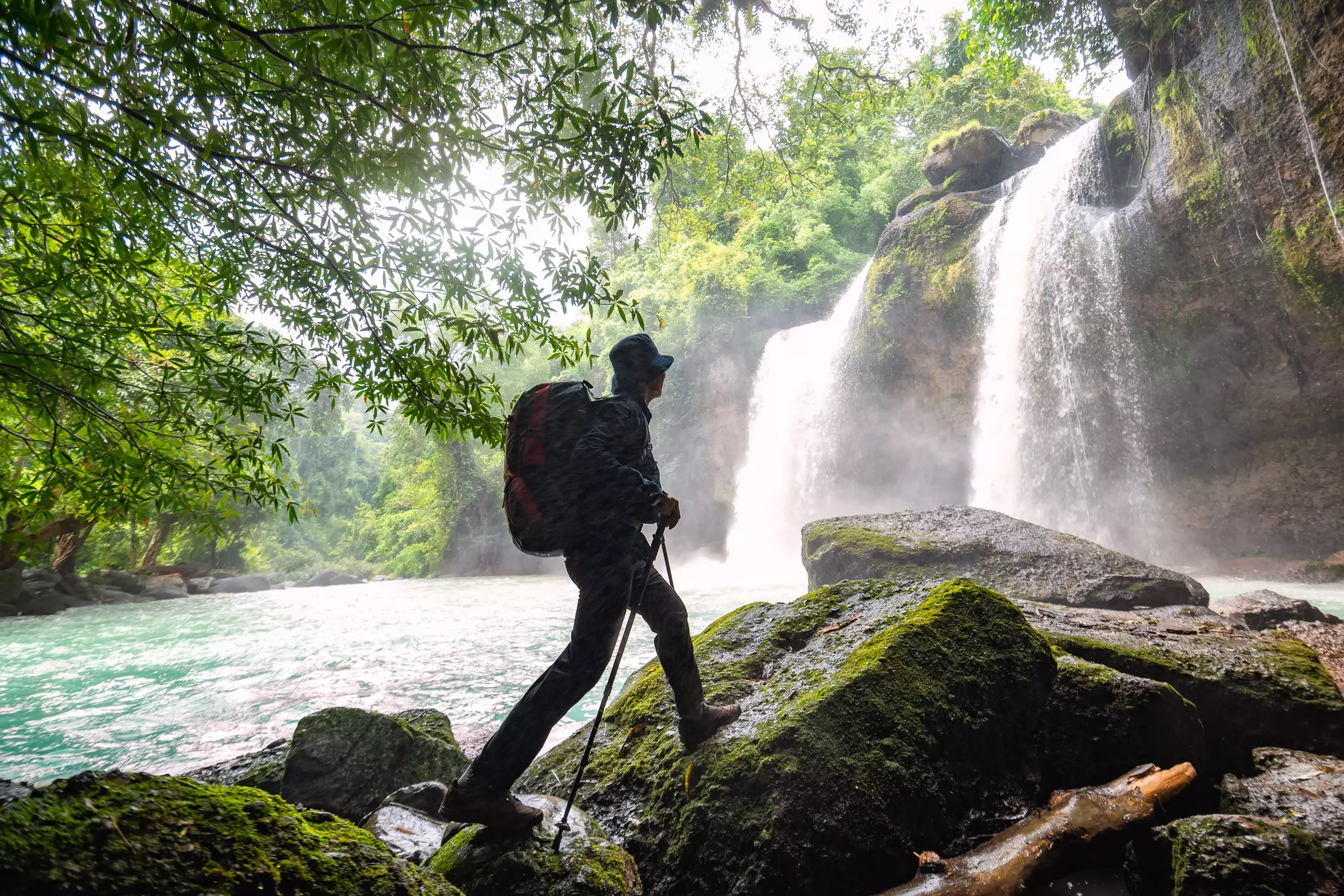 A hiker with poles and a large backpack is in silhouette walking on mossy rocks in front of a tall waterfall in a rainforest in Thailand.