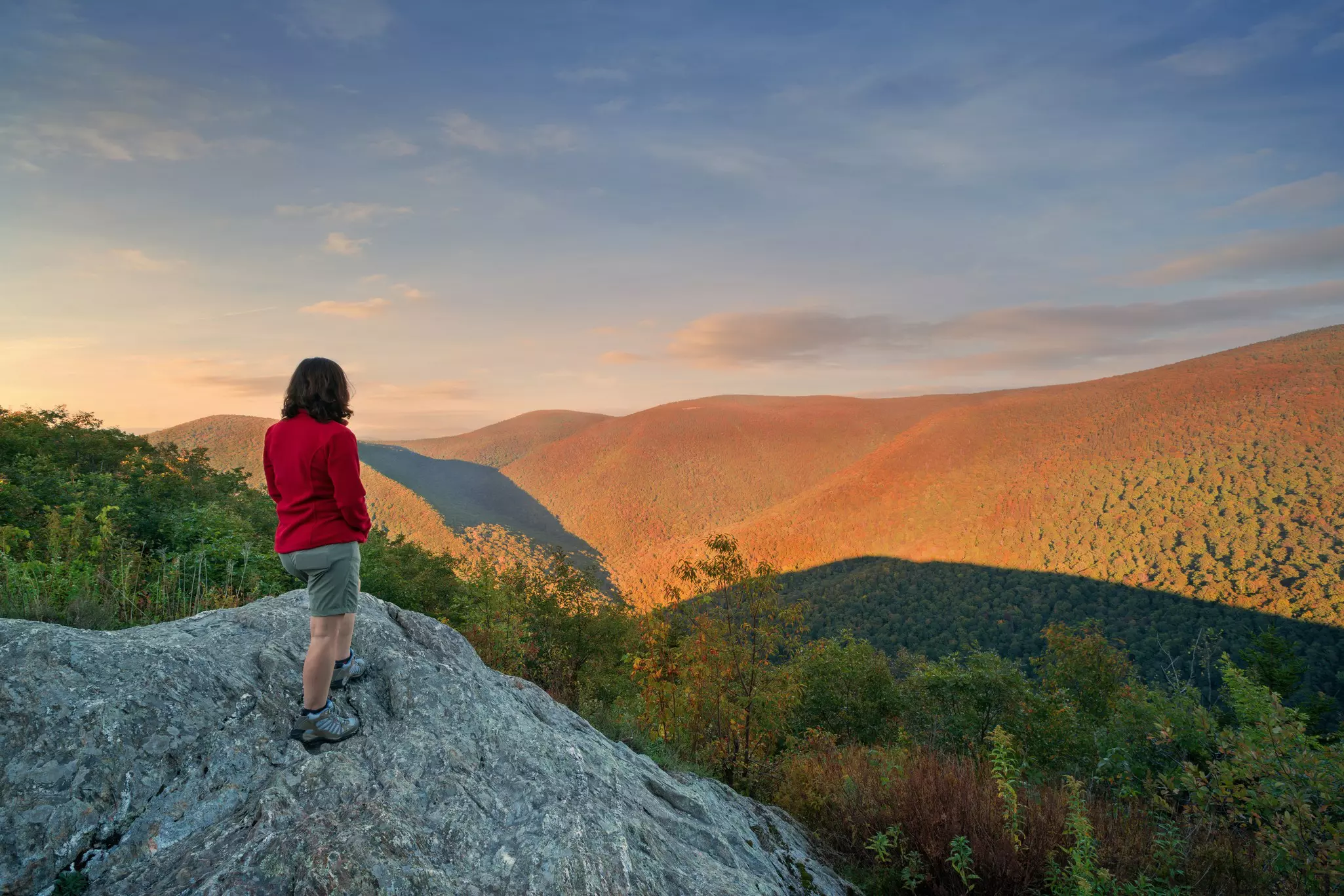 A hiker in a red shirt stands on a rock looking at mountains with fall foliage in early evening.
