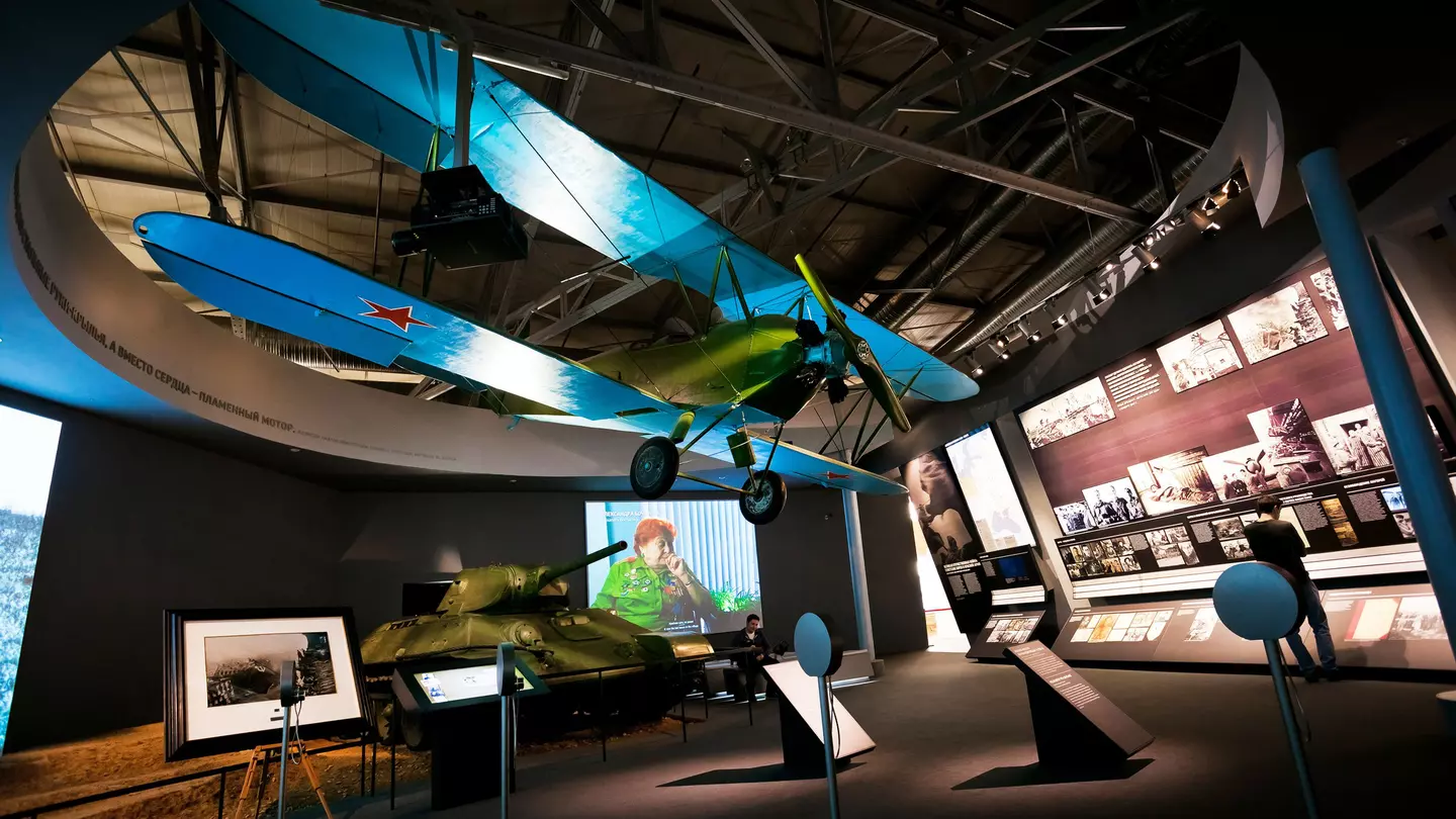 A large dark room, with many illuminated exhibits including a wall of black-and-white photographs. A tank stands against a wall and a biplane hangs from the ceiling