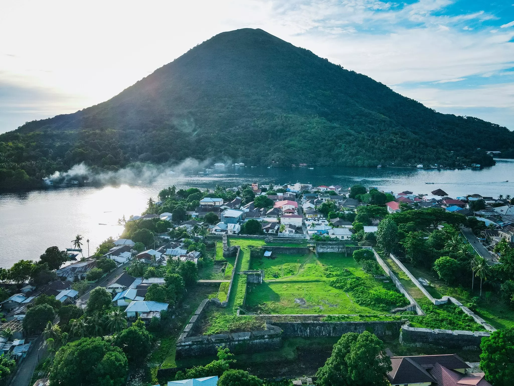 An aerial view of the outline of a former fortress now covered in grass, next to a village and beside a channel between hilly green islands.