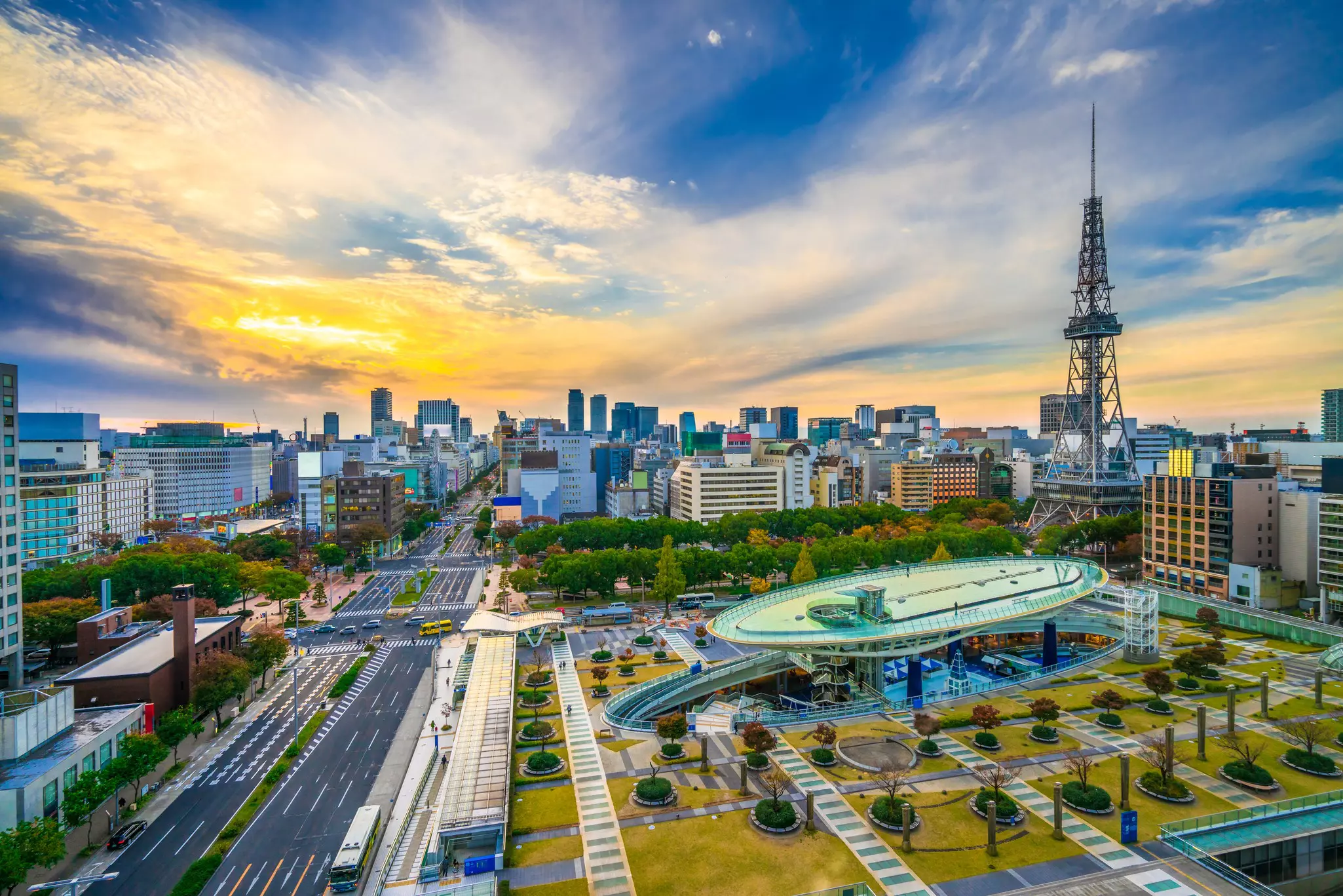 A city skyline at sunset. A large space is dominated by a dish-like piece of architecture overlooked by a tall communications tower.