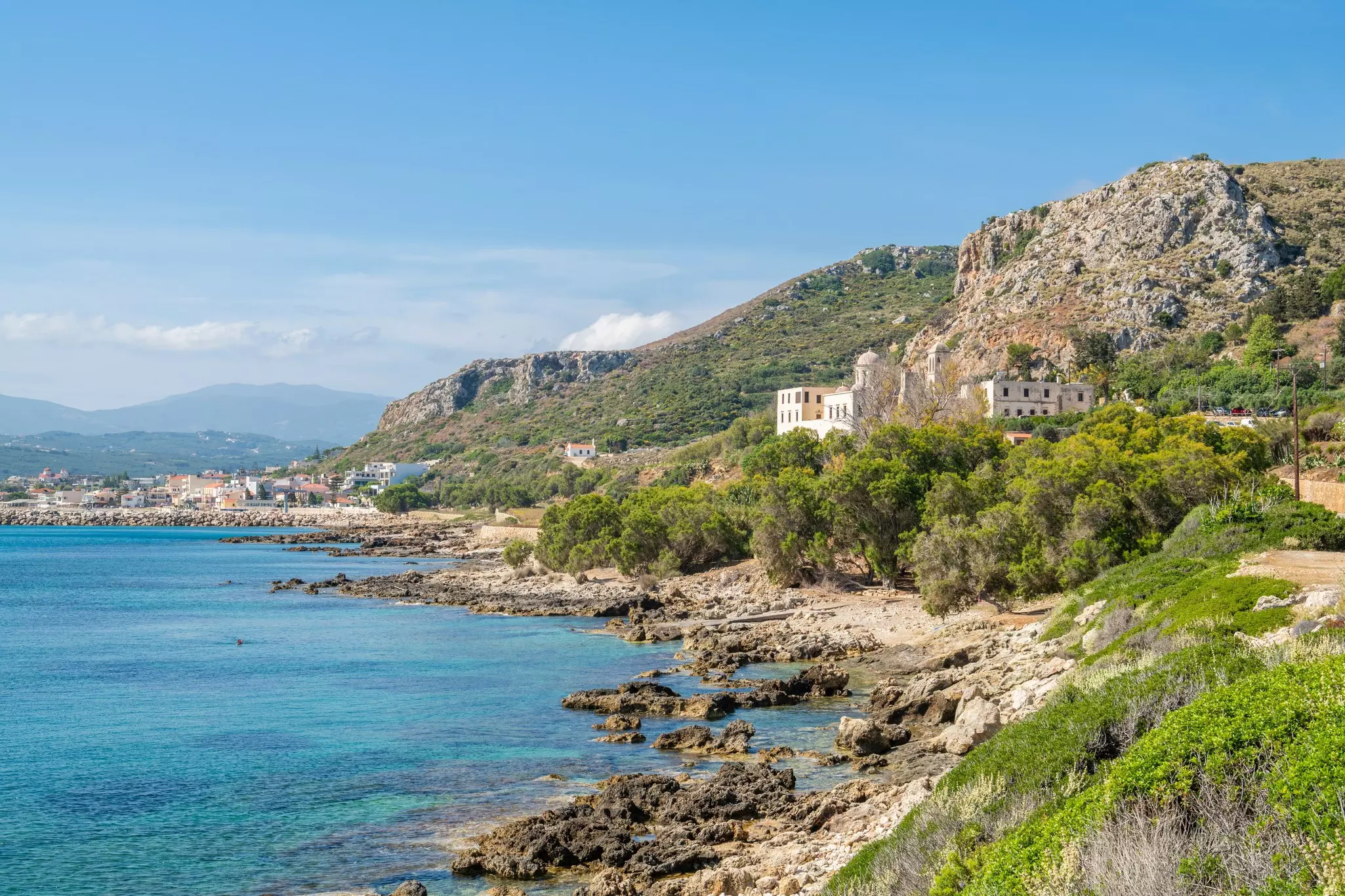 A view of a rocky coast, with buildings on a hillside and cliffs in the distance.
