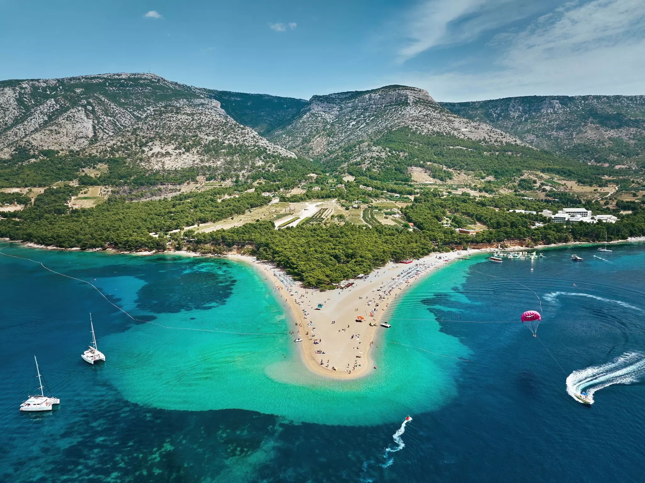 An aerial view of a beach on a headland, surrounded by blue-green waters, with mountains in the distance.