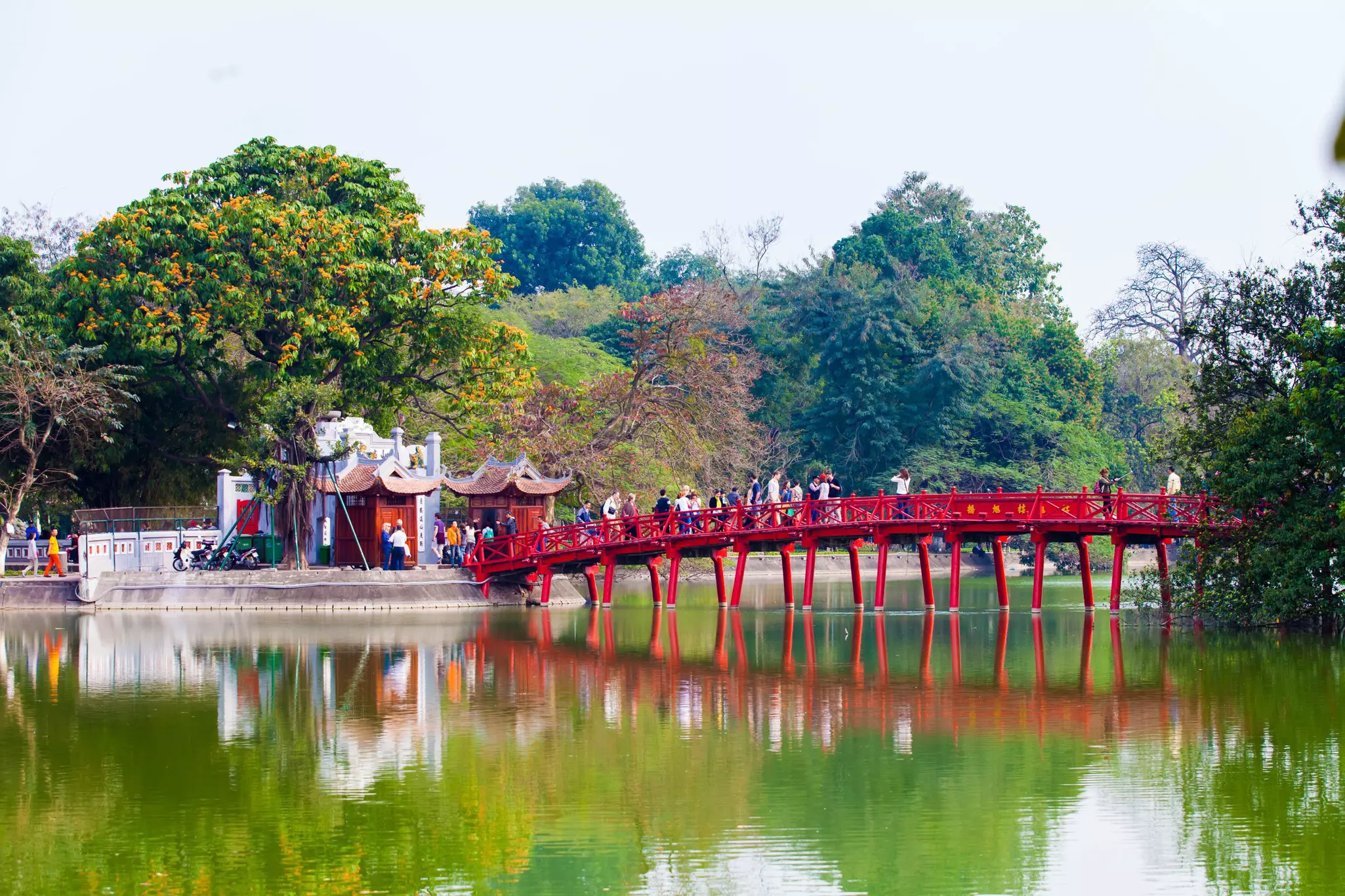 People walk over a red bridge leading to a temple on a lake.
