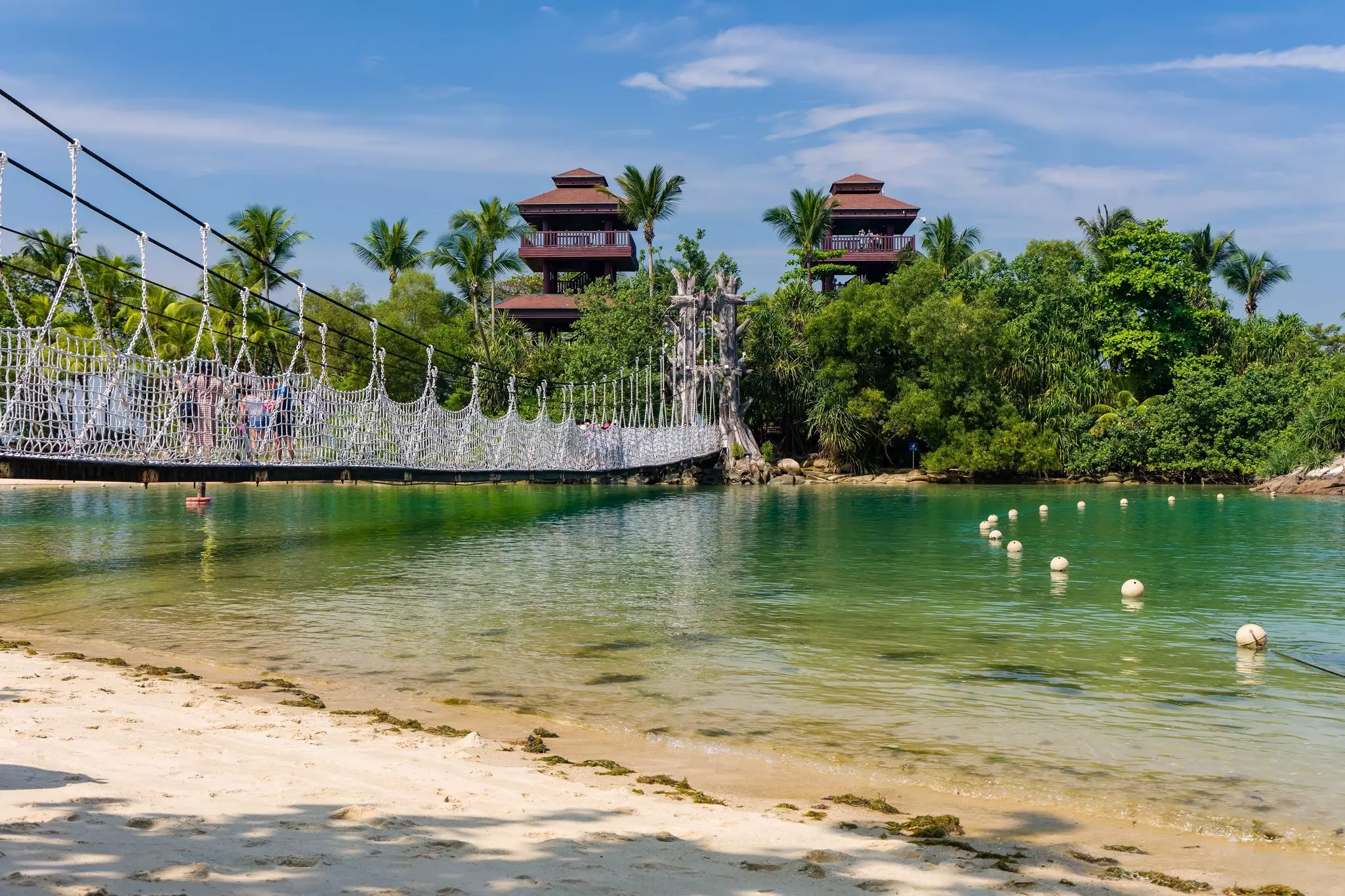 A suspension bridge just above the water level connects a beach and a small island with two wooden lookout towers.