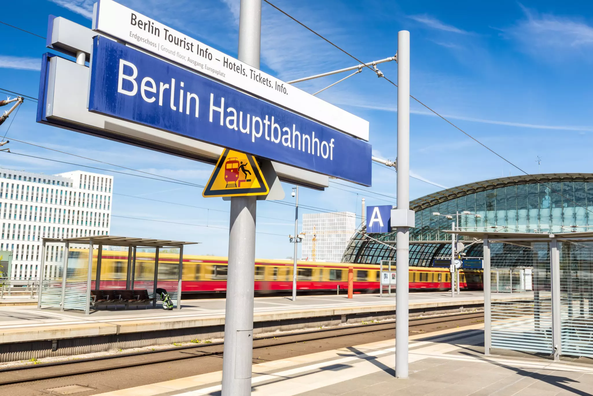 Train station platform with red and yellow train speeding past in the background and blue sign reading "Berlin Hauptbahnhof" in the foreground on a sunny day.